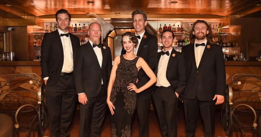 Ashley Anne in a beaded flapper gown and headpiece with The Carnations in black tie at a warmly lit vintage speakeasy bar